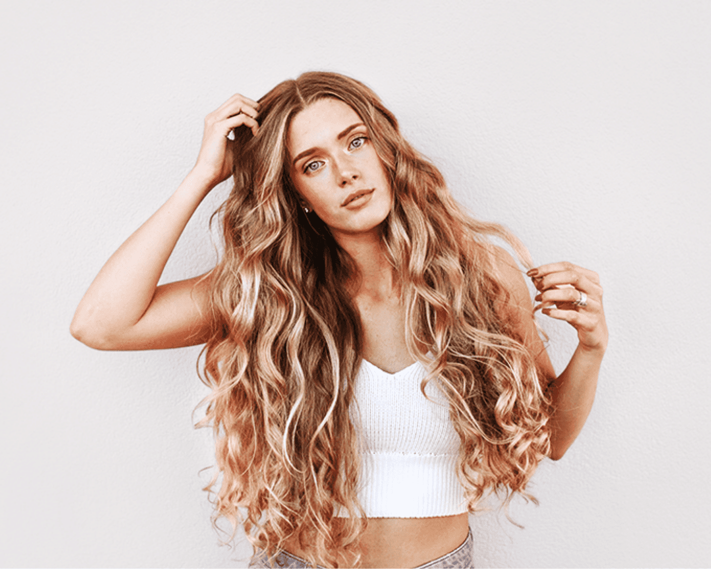 Young woman with long wavy hair, wearing a white top, posing against a plain background.